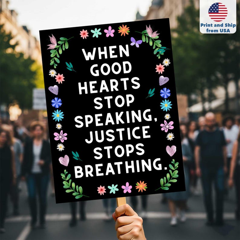 May include: A black sign with the text "WHEN GOOD HEARTS STOP SPEAKING, JUSTICE STOPS BREATHING." is held up in a crowd. The sign is decorated with colorful flowers and leaves. The upper right corner has a small American flag.