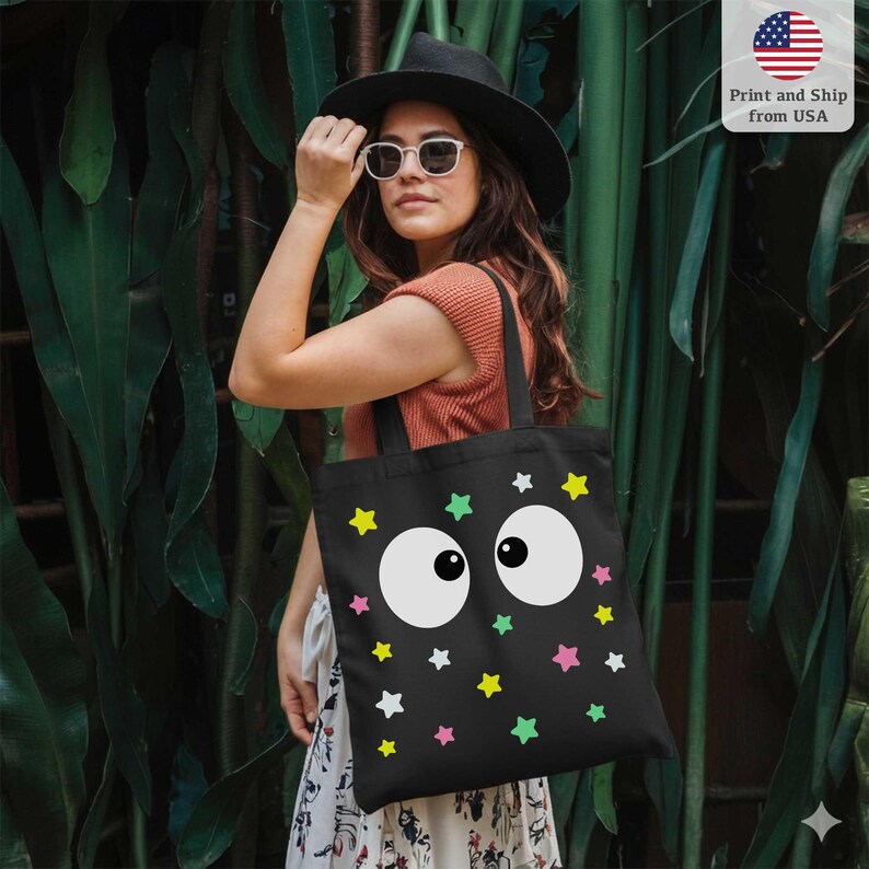 Puede incluir: Bolso tote negro con un dise&ntilde;o caprichoso de grandes ojos blancos de dibujos animados y estrellas de colores en amarillo, verde y rosa. El bolso tiene asas negras. El fondo es un exuberante follaje verde.