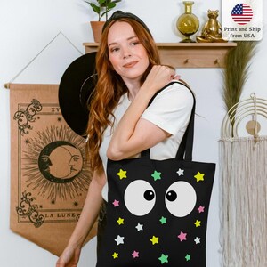 Puede incluir: Bolso tote negro con un dise&ntilde;o caprichoso de grandes ojos blancos de dibujos animados y estrellas de colores. El bolso lo lleva una persona. El fondo incluye una pared con decoraci&oacute;n.