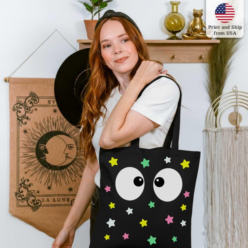 Puede incluir: Bolso tote negro con un dise&ntilde;o caprichoso de grandes ojos blancos de dibujos animados y estrellas de colores. El bolso lo lleva una persona. El fondo incluye una pared con decoraci&oacute;n.