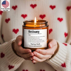 Puede incluir: Una vela encendida en un tarro de cristal con la etiqueta "Bethany". La etiqueta también incluye una definición y la fragancia "Frambuesa & Vainilla". La vela se sostiene con ambas manos.