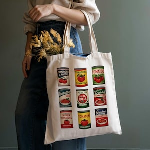May include: A white tote bag featuring colorful illustrations of tomato cans. The cans display various brands and styles, including "Pomodoro di Napoli," "Tomatoo," and "Crushed Tomatoes." The bag is held by a person.