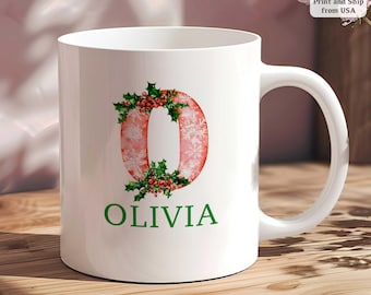 Op de afbeelding: Witte keramische mok met een grote letter "O" versierd met sneeuwvlokken en hulst. De letter is roze, met de naam "OLIVIA" in groen gedrukt. Een feestelijk, gepersonaliseerd cadeau.