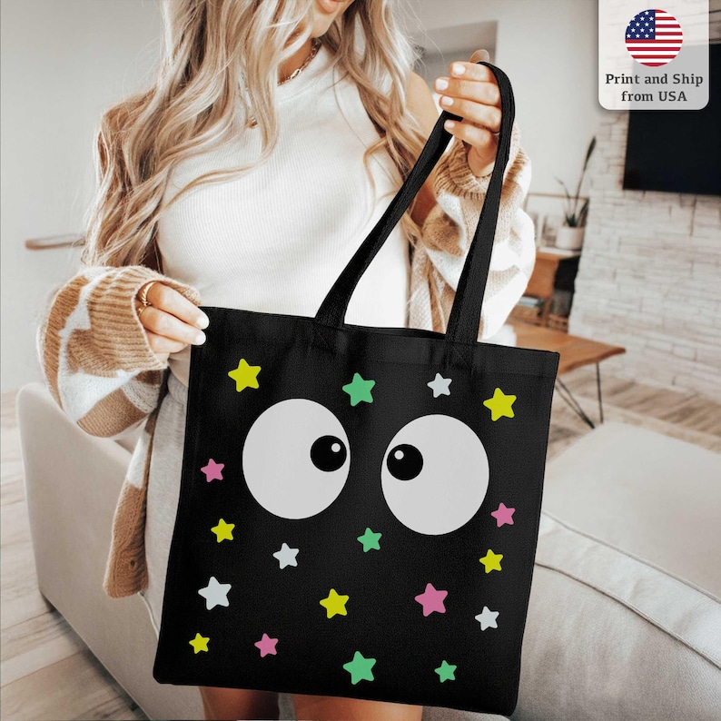 Puede incluir: Bolso tote negro con un dise&ntilde;o caprichoso de grandes ojos blancos de dibujos animados y estrellas de colores en amarillo, verde, rosa y blanco. Tiene asas largas negras.