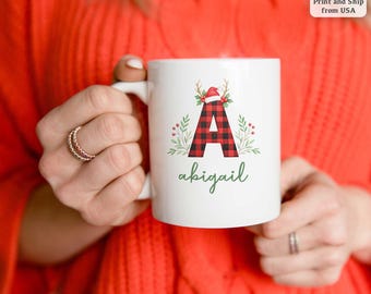 Op de afbeelding: Witte keramische mok met een rood-zwart geruit letter "A" ontwerp, versierd met een kerstmuts en geweien. De naam "abigail" staat eronder in groen schrift. De mok wordt vastgehouden door een persoon met ringen.