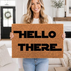 Pode incluir: Um capacho retangular de fibra de coco castanha com o texto HELLO THERE em letras maiúsculas pretas e em negrito. O tapete tem uma textura fibrosa e áspera, com um tipo de letra moderno sem serifa centrado na parte frontal.