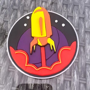 May include: A circular patch featuring a stylized rocket ship design. The rocket is yellow and orange, set against a purple background with a black border. The patch has a red and white outer edge, with white stars.