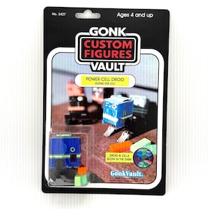 Op de afbeelding: Een verpakte Power Cell Droid speelfiguur van Gonk Custom Figures Vault. Het blauwe en oranje speelgoed zit in een doorzichtige plastic hoes op een zwarte achtergrond. De verpakking bevat de tekst "Ages 4 and up" en "DROID & CELLS GLOW IN THE DARK."