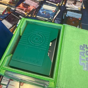 May include: A green Star Wars card storage box with a matching green interior. The box is open, revealing a stack of cards and a green card holder. Several other cards are scattered around the box on a dark blue surface.