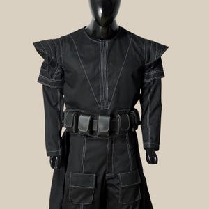 May include: A black costume with white stitching, featuring a long-sleeved top with shoulder accents and a matching skirt. The outfit includes a belt with pouches and is displayed on a mannequin.