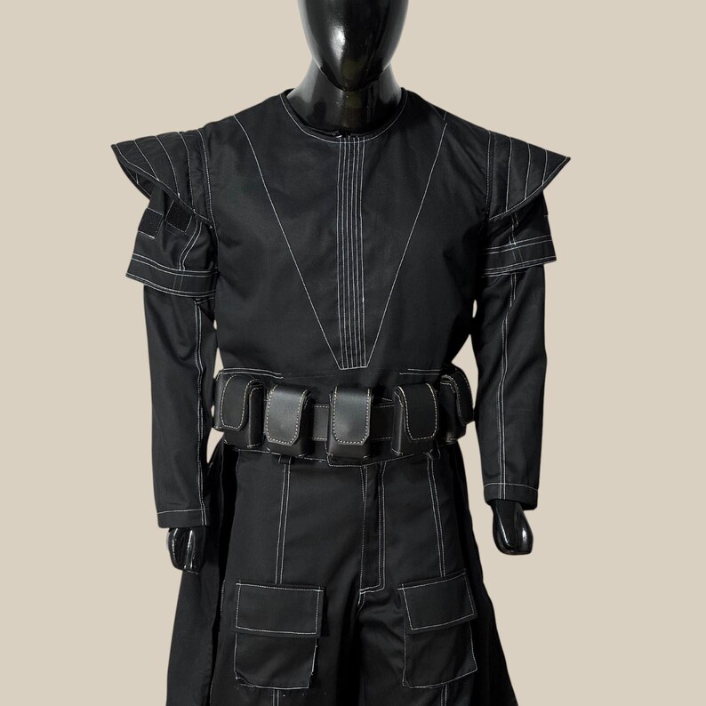 May include: A black costume with white stitching, featuring a long-sleeved top with shoulder accents and a matching skirt. The outfit includes a belt with pouches and is displayed on a mannequin.