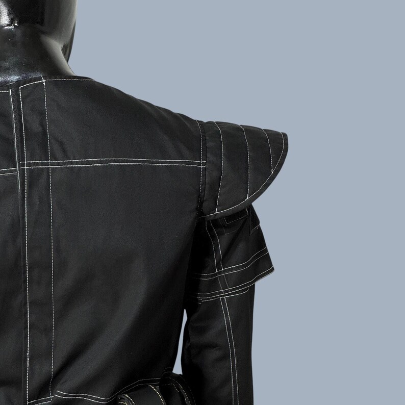 May include: Black jacket with white stitching details. The jacket features structured shoulders and a unique design. The stitching creates a geometric pattern across the garment. The jacket is made of a smooth, possibly synthetic material.