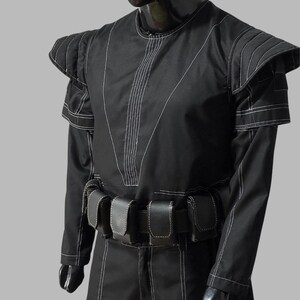 May include: Black costume with white stitching details. The outfit features a round neck, structured shoulders, and a belt with multiple pouches. The sleeves are long, and the stitching creates a geometric pattern. The costume is displayed on a mannequin.