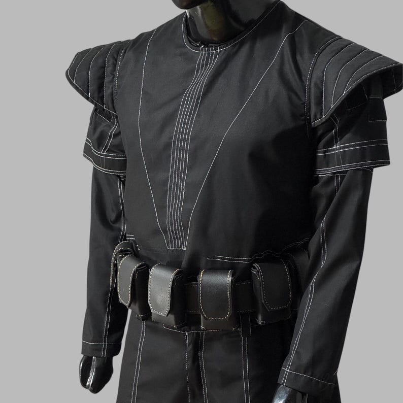 May include: Black costume with white stitching details. The outfit features a round neck, structured shoulders, and a belt with multiple pouches. The sleeves are long, and the stitching creates a geometric pattern. The costume is displayed on a mannequin.