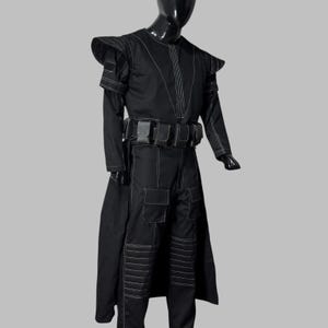 May include: Black costume with white stitching, displayed on a mannequin. The outfit includes a long-sleeved top with shoulder accents, a belt with pouches, and matching pants. The costume has a long coat-like element in the back.