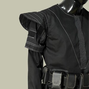 May include: Black garment with white stitching details. The garment features a structured shoulder design with a flap and a velcro patch. A series of black pouches are visible at the waist. The outfit is displayed on a black mannequin.
