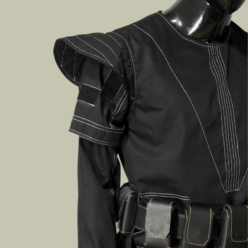 May include: Black garment with white stitching details. The garment features a structured shoulder design with a flap and a velcro patch. A series of black pouches are visible at the waist. The outfit is displayed on a black mannequin.