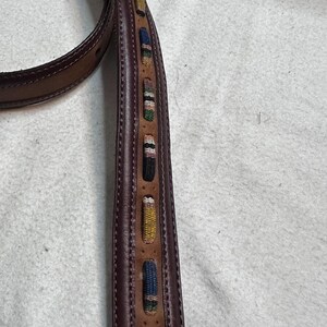 K&ouml;nnte beinhalten: Brauner Lederg&uuml;rtel mit dekorativen gewebten Akzenten in verschiedenen Farben. Der G&uuml;rtel hat ein gen&auml;htes Design und eine klassische Schnalle. Die gewebten Details umfassen blaue, gelbe, gr&uuml;ne und rosa Muster.