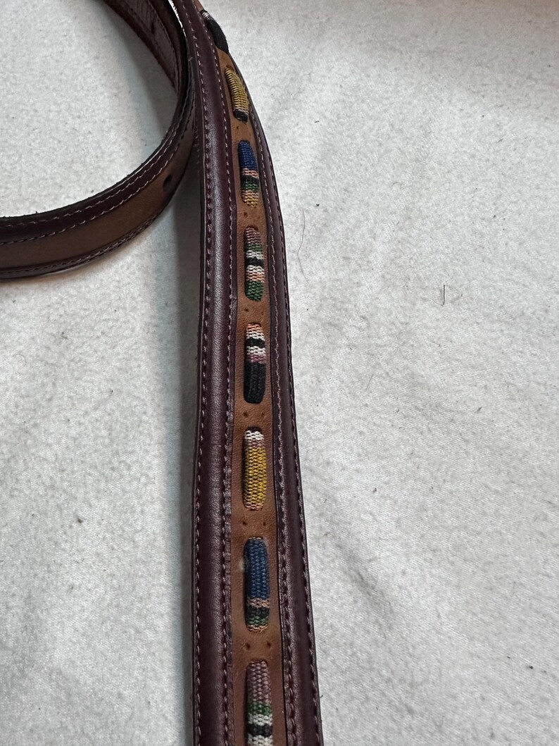 K&ouml;nnte beinhalten: Brauner Lederg&uuml;rtel mit dekorativen gewebten Akzenten in verschiedenen Farben. Der G&uuml;rtel hat ein gen&auml;htes Design und eine klassische Schnalle. Die gewebten Details umfassen blaue, gelbe, gr&uuml;ne und rosa Muster.