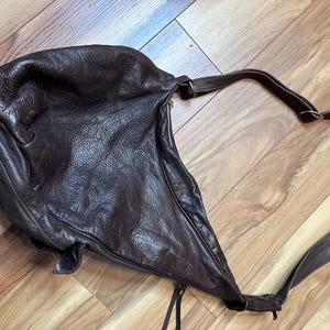 May include: A dark brown leather shoulder bag with a long strap. The bag has a slouchy, unstructured design and a zipper closure. The leather appears to be textured and worn, giving it a vintage look.