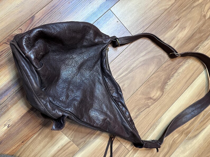 May include: A dark brown leather shoulder bag with a long strap. The bag has a slouchy, unstructured design and a zipper closure. The leather appears to be textured and worn, giving it a vintage look.