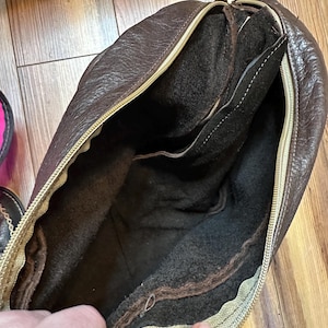 May include: A brown leather bag with a gold zipper, open to reveal a dark brown interior. The bag has a textured exterior and a soft, dark interior lining. The zipper is a contrasting gold color.