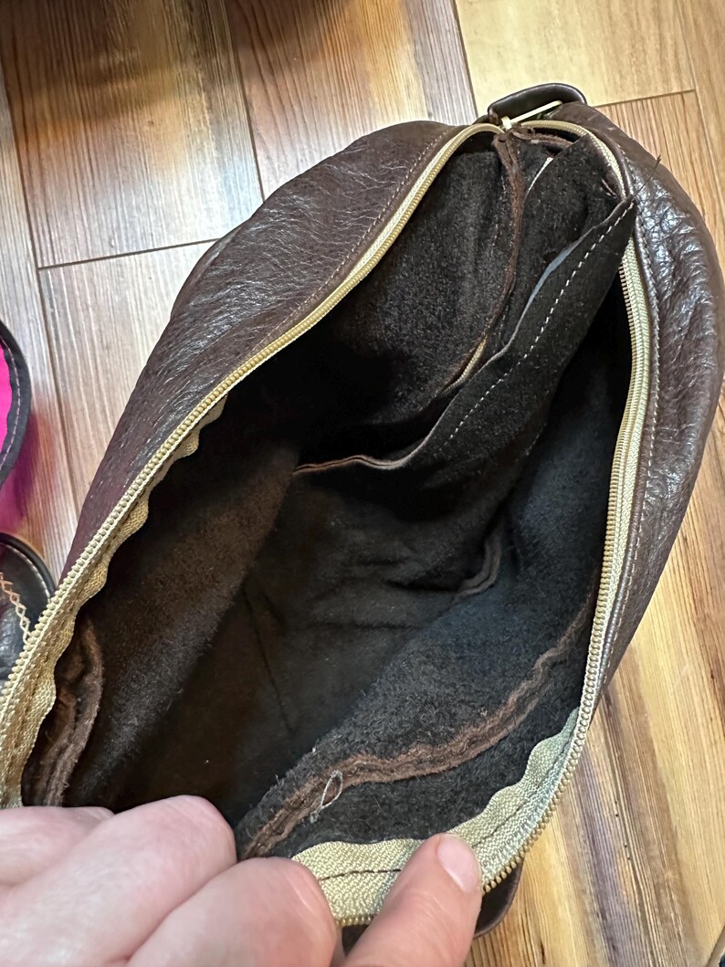 May include: A brown leather bag with a gold zipper, open to reveal a dark brown interior. The bag has a textured exterior and a soft, dark interior lining. The zipper is a contrasting gold color.