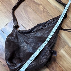 May include: A dark brown leather crossbody bag with a long adjustable strap. The bag is measured with a tape measure, showing a length of approximately 12 inches. The leather has a textured, wrinkled appearance.