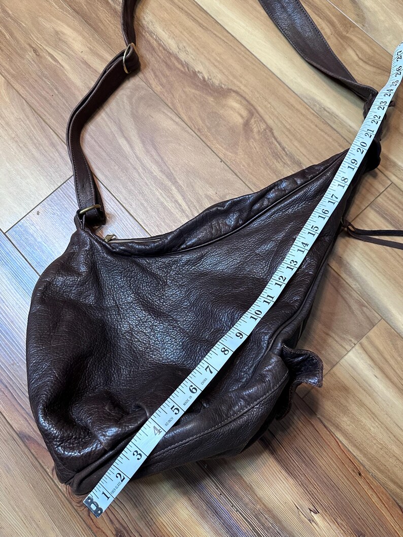May include: A dark brown leather crossbody bag with a long adjustable strap. The bag is measured with a tape measure, showing a length of approximately 12 inches. The leather has a textured, wrinkled appearance.