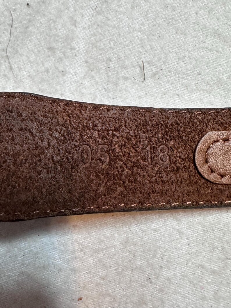 K&ouml;nnte beinhalten: Nahaufnahme eines braunen Lederg&uuml;rtels mit den gepr&auml;gten Zahlen "05 18". Der G&uuml;rtel hat eine strukturierte Oberfl&auml;che und einen gen&auml;hten Rand. Die Schnalle ist auf der rechten Seite sichtbar.