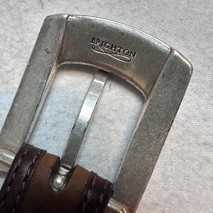 K&ouml;nnte beinhalten: Nahaufnahme einer silberfarbenen G&uuml;rtelschnalle mit dem eingravierten Markennamen "BRIGHTON". Die Schnalle ist an einem braunen Lederg&uuml;rtel mit dunklen N&auml;hten befestigt.