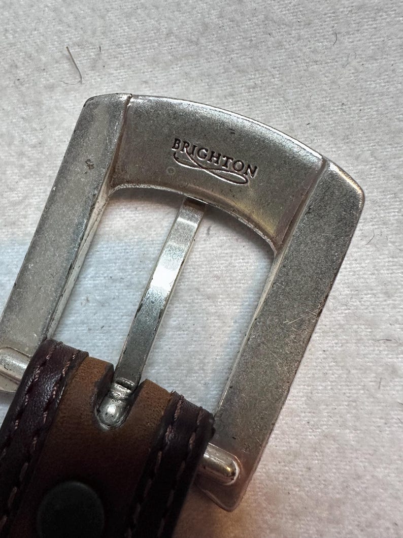K&ouml;nnte beinhalten: Nahaufnahme einer silberfarbenen G&uuml;rtelschnalle mit dem eingravierten Markennamen "BRIGHTON". Die Schnalle ist an einem braunen Lederg&uuml;rtel mit dunklen N&auml;hten befestigt.