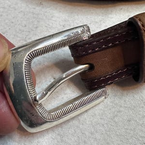 K&ouml;nnte beinhalten: Nahaufnahme eines braunen Lederg&uuml;rtels mit einer silberfarbenen Schnalle. Die rechteckige Schnalle hat ein strukturiertes Design. Der G&uuml;rtel hat einen gen&auml;hten Rand und ein kontrastierendes dunkelbraunes Innenfutter.