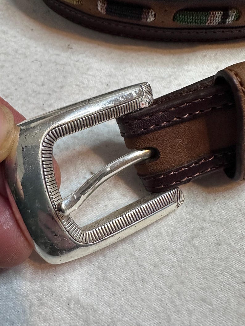 K&ouml;nnte beinhalten: Nahaufnahme eines braunen Lederg&uuml;rtels mit einer silberfarbenen Schnalle. Die rechteckige Schnalle hat ein strukturiertes Design. Der G&uuml;rtel hat einen gen&auml;hten Rand und ein kontrastierendes dunkelbraunes Innenfutter.