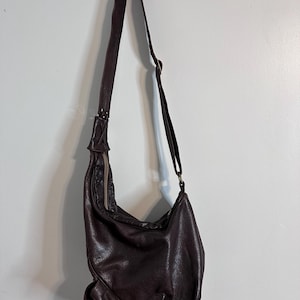 May include: A dark brown leather shoulder bag with a long adjustable strap. The bag has a slouchy design with a zipper closure and a textured surface. The bag is hanging against a white wall.