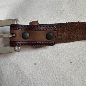 K&ouml;nnte beinhalten: Nahaufnahme eines braunen Lederg&uuml;rtels mit einer silbernen Schnalle. Der G&uuml;rtel hat einen dunkelbraunen Lederriemen mit einem Nahtmuster und zwei schwarzen Nieten in der N&auml;he der Schnalle. Die Schnalle ist silberfarben gl&auml;nzend.