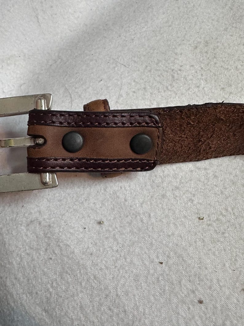 K&ouml;nnte beinhalten: Nahaufnahme eines braunen Lederg&uuml;rtels mit einer silbernen Schnalle. Der G&uuml;rtel hat einen dunkelbraunen Lederriemen mit einem Nahtmuster und zwei schwarzen Nieten in der N&auml;he der Schnalle. Die Schnalle ist silberfarben gl&auml;nzend.