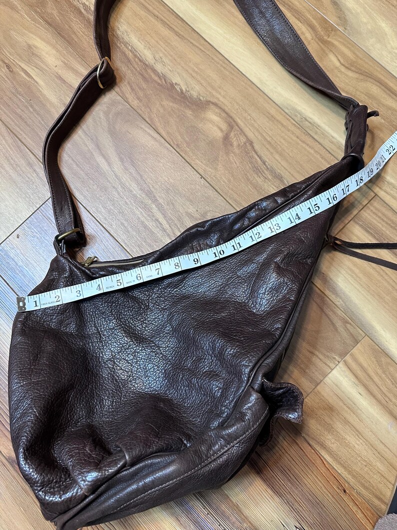 May include: A dark brown leather shoulder bag with a long strap. The bag is measured with a tape measure, showing a length of approximately 22 inches. The leather has a textured appearance.