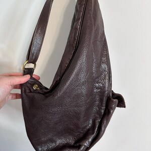 May include: A dark brown leather shoulder bag with a long strap and a rounded shape. The bag has a textured surface and a brass-colored metal ring and clasp. The strap is attached to the bag with a brass-colored metal ring.