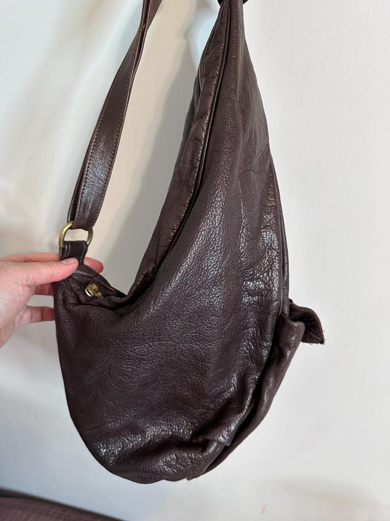 May include: A dark brown leather shoulder bag with a long strap and a rounded shape. The bag has a textured surface and a brass-colored metal ring and clasp. The strap is attached to the bag with a brass-colored metal ring.