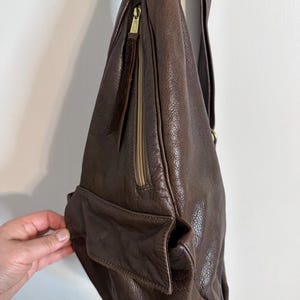 May include: A dark brown leather shoulder bag with a long strap. The bag features a zippered pocket with a gold-colored zipper and a small exterior pocket. The leather has a textured appearance.