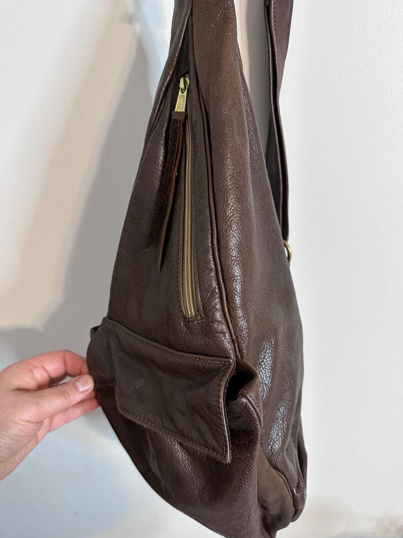 May include: A dark brown leather shoulder bag with a long strap. The bag features a zippered pocket with a gold-colored zipper and a small exterior pocket. The leather has a textured appearance.