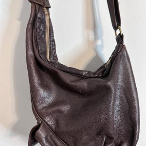 May include: A dark brown leather shoulder bag with a long strap and a zipper closure. The bag has a textured surface and a braided detail at the top. The strap is attached to the bag with a gold-colored ring.