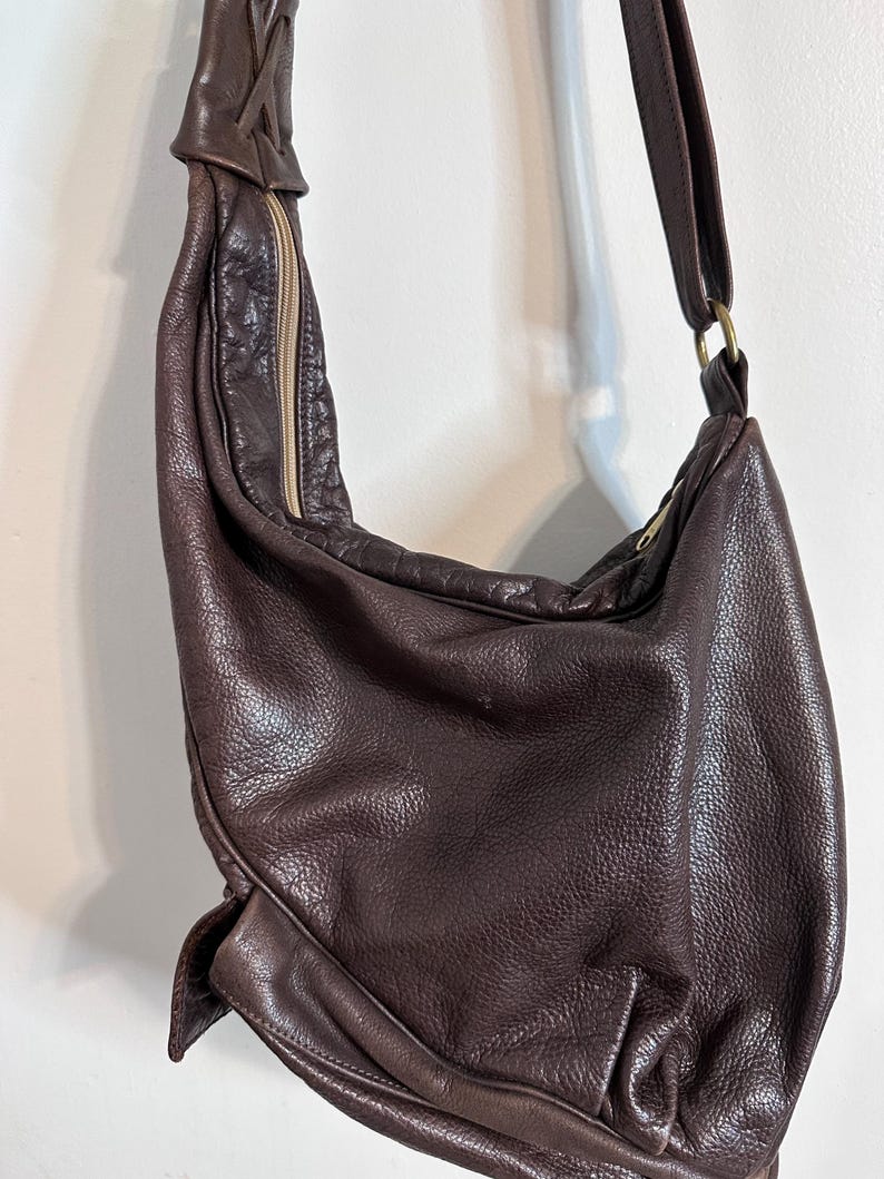 May include: A dark brown leather shoulder bag with a long strap and a zipper closure. The bag has a textured surface and a braided detail at the top. The strap is attached to the bag with a gold-colored ring.
