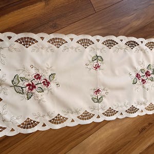 May include: Cream-colored embroidered table runner with a scalloped edge. Features floral embroidery in shades of burgundy, white, and green. The runner is approximately 30 inches long and is placed on a wooden surface.