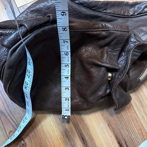 May include: A dark brown leather shoulder bag with a soft, textured appearance. The bag is measured with a tape measure, showing a width of approximately 6 inches. The bag has a zipper and a strap.