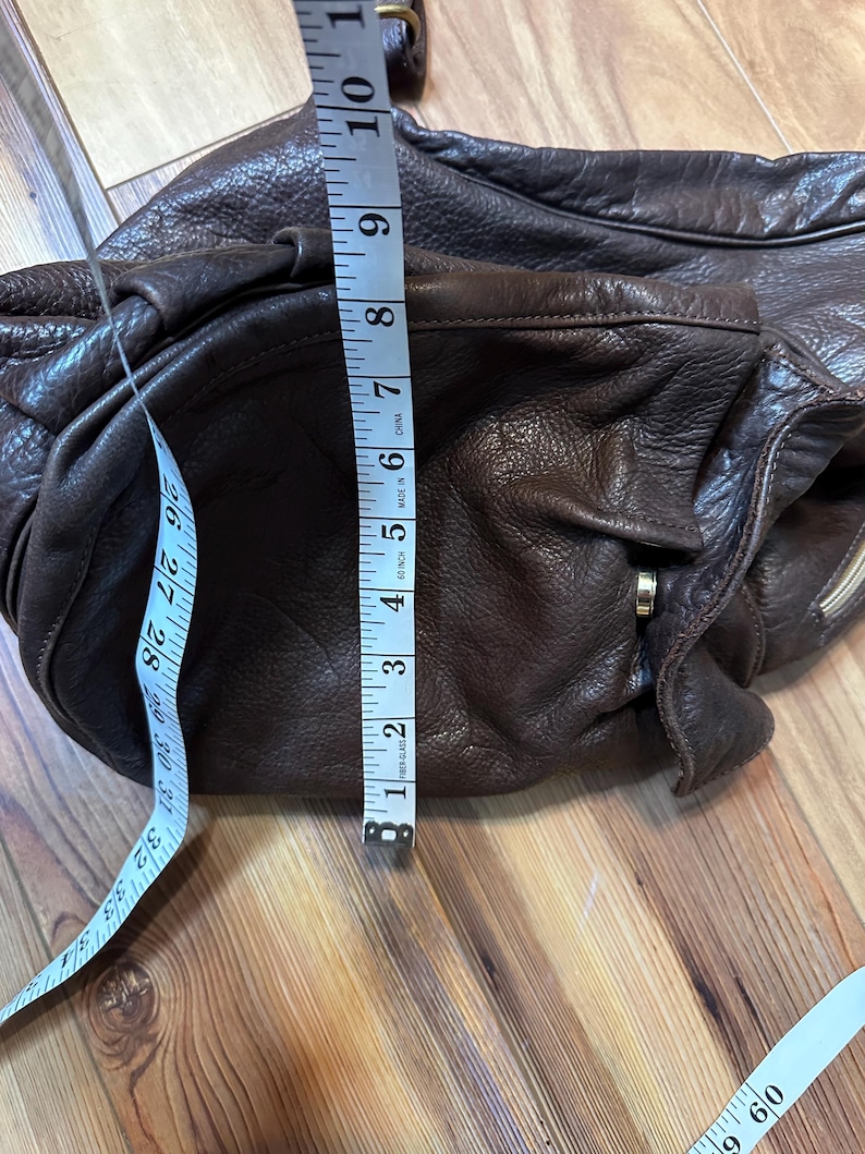 May include: A dark brown leather shoulder bag with a soft, textured appearance. The bag is measured with a tape measure, showing a width of approximately 6 inches. The bag has a zipper and a strap.