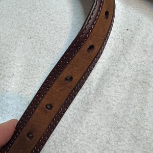 K&ouml;nnte beinhalten: Nahaufnahme eines braunen Lederg&uuml;rtels mit dunkelburgunderfarbenem Rand und N&auml;hten. Der G&uuml;rtel hat mehrere gleichm&auml;&szlig;ig verteilte L&ouml;cher und eine hellere braune Mitte. Das Leder scheint von hoher Qualit&auml;t zu sein.