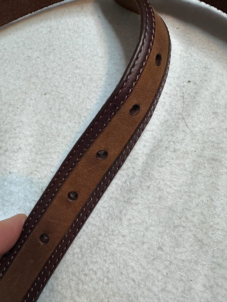 K&ouml;nnte beinhalten: Nahaufnahme eines braunen Lederg&uuml;rtels mit dunkelburgunderfarbenem Rand und N&auml;hten. Der G&uuml;rtel hat mehrere gleichm&auml;&szlig;ig verteilte L&ouml;cher und eine hellere braune Mitte. Das Leder scheint von hoher Qualit&auml;t zu sein.