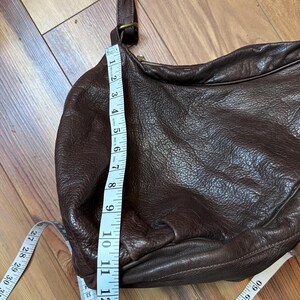 May include: A brown leather crossbody bag with a long adjustable strap. The bag is measured at approximately 13 inches in height. The bag has a zipper closure and a rounded shape. The bag is made of a textured leather material.
